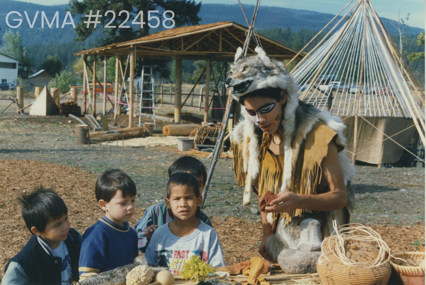 An outdoor camp setup. In the background is a wooden roofed structure, as well as a teepee. In the foreground is four children and an adult. The four little boys have dark hair. Three are watching the adult, while one is looking at the camera. The adult is wearing leather clothing, and his face is panted in black and white. He has a wolf skin over his head and hanging down his shoulders. He is looking down at a rock in his head. In front of him are two woven baskets.