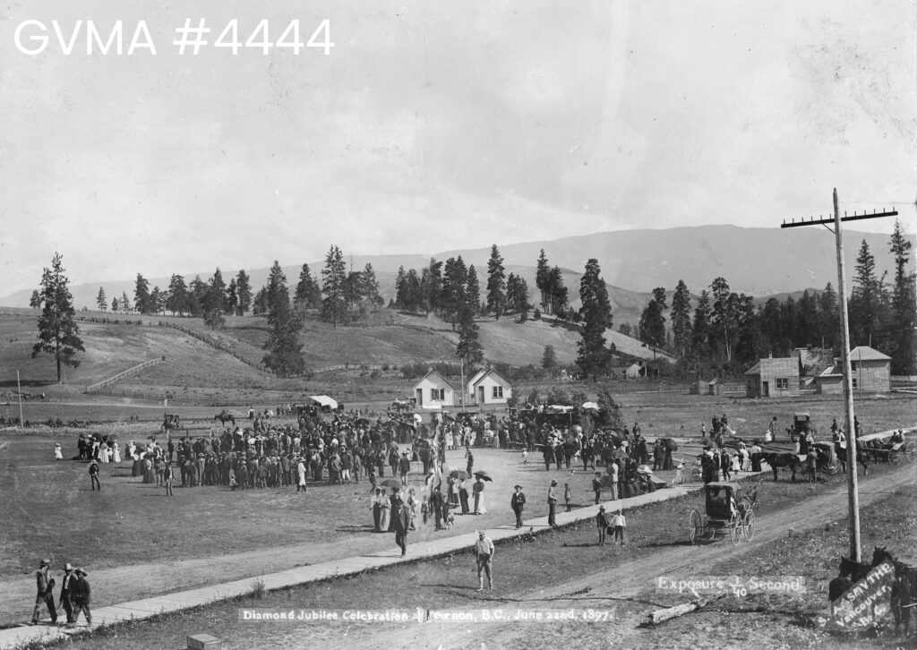 Crowds of people walking a long a board walk, some with parasols in a park. Some structures are visible in the background, and a few horses and buggies along the right side. A hillside with trees is visible in the background.