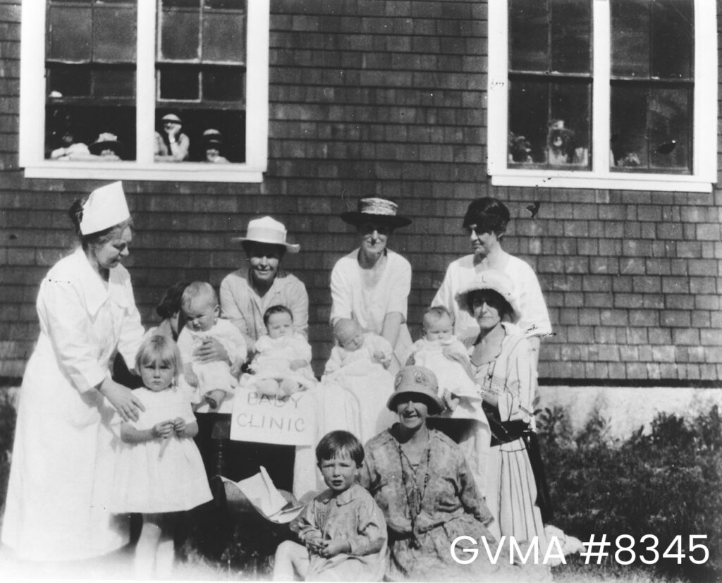 A black and white photo of women and children in front of a wooden structure. A sign in between the people reads "Baby Clinic." A nurse is standing to the left with her hands on the shoulders of a little girl. Women in hands holding babies are to the right. Several people are looking out from two windows on the building in the background.