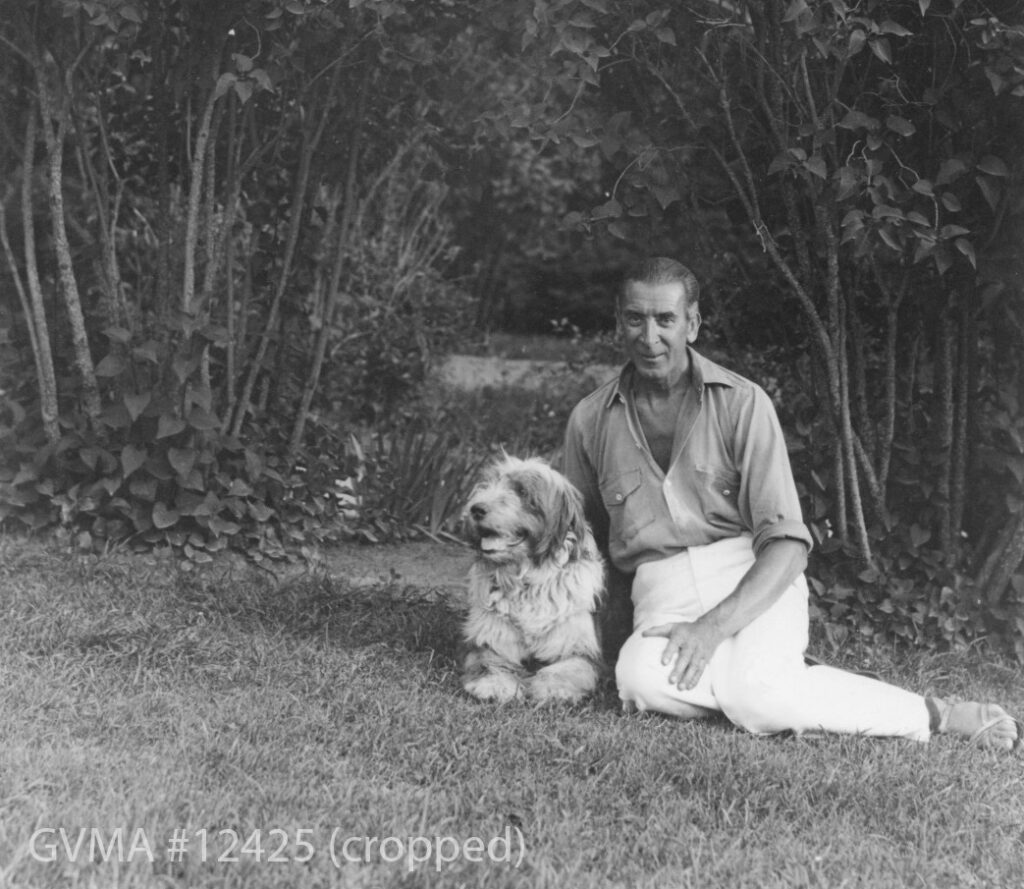 A black and white photo of a man lounging on the lawn with a dog. He has short, slicked-back hair, and is wearing a loose shirt, unbuttoned beneath the collar bone, and white pants. He has sandals on his feet. The dog has his mouth open with his tongue out and is looking away from the camera. The photo is framed in the background by overhanding bushes.