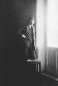 A black and white photo of a man standing, with one knee up on chair. He is positioned near a window through which light is streaming. He has one hand on his hip, and is wearing a bowler hat.