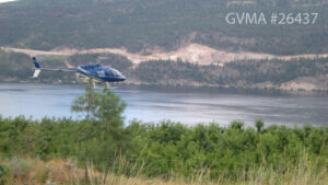 A blue helicopter flying over green cherry trees, In the background is a lake, and behind that a mountain with trees and several clear cuttings. 