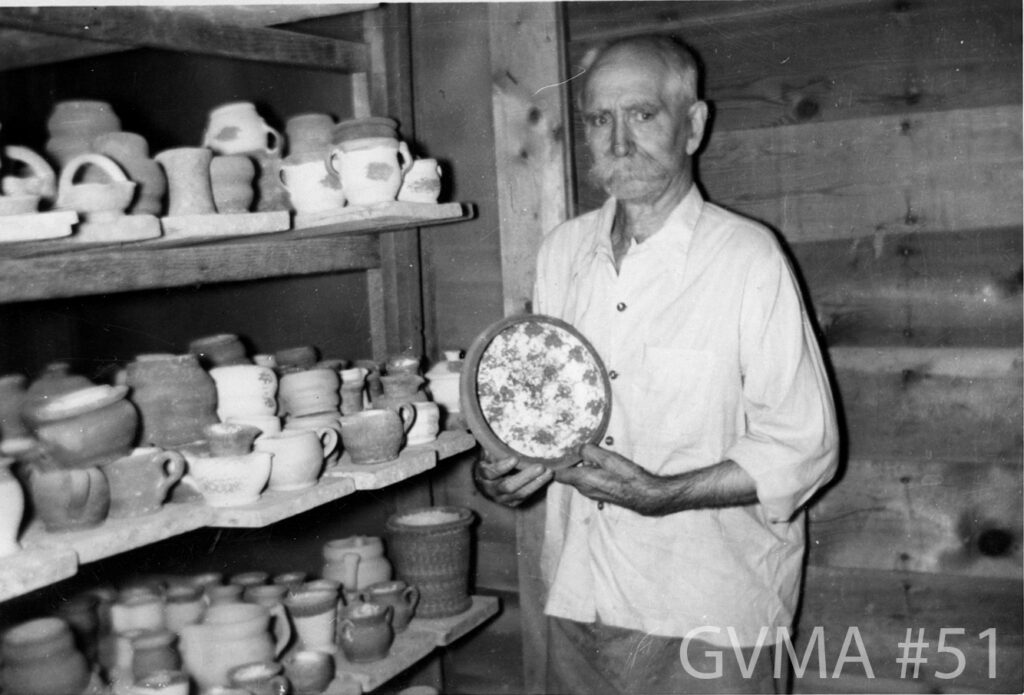 A man in black and white wearing a loose, white button-up shirt standing in a pottery studio. He has a bushy mustache and furrowed eyebrows, and a partially bald head. He is showing a pottery bowl to the camera. To his left are three shelved full of different pieces of pottery, including bowls, mugs, and jugs.