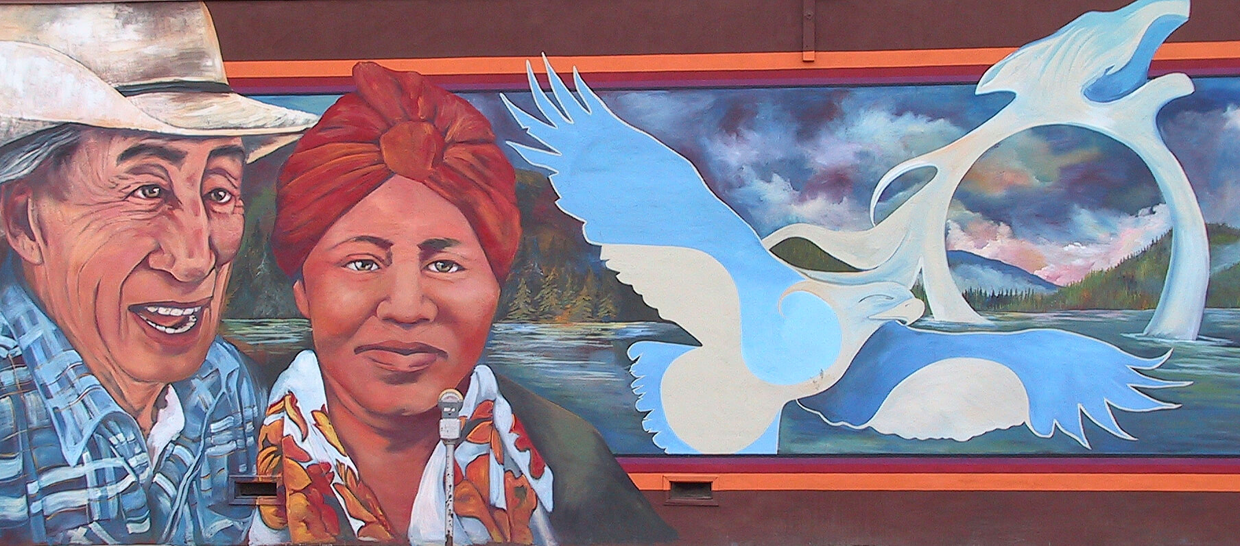 Vernon's Okanagan Indian Band Mural. An older indigenous man with gray hair wearing a cowboy hat is located on the left of the image. He is smiling and wearing a blue plaid shirt. Next to him is a woman, smiling slightly, and wearing an orange head scarf, and a white and orange scarf around her neck. To her right is a blue stylized eagle, wolf, and sea serpent design. The background shows a blue, multicoloured lake, with green forests and blue mountains in the background. The blue-black sky is doted with pink clouds. 