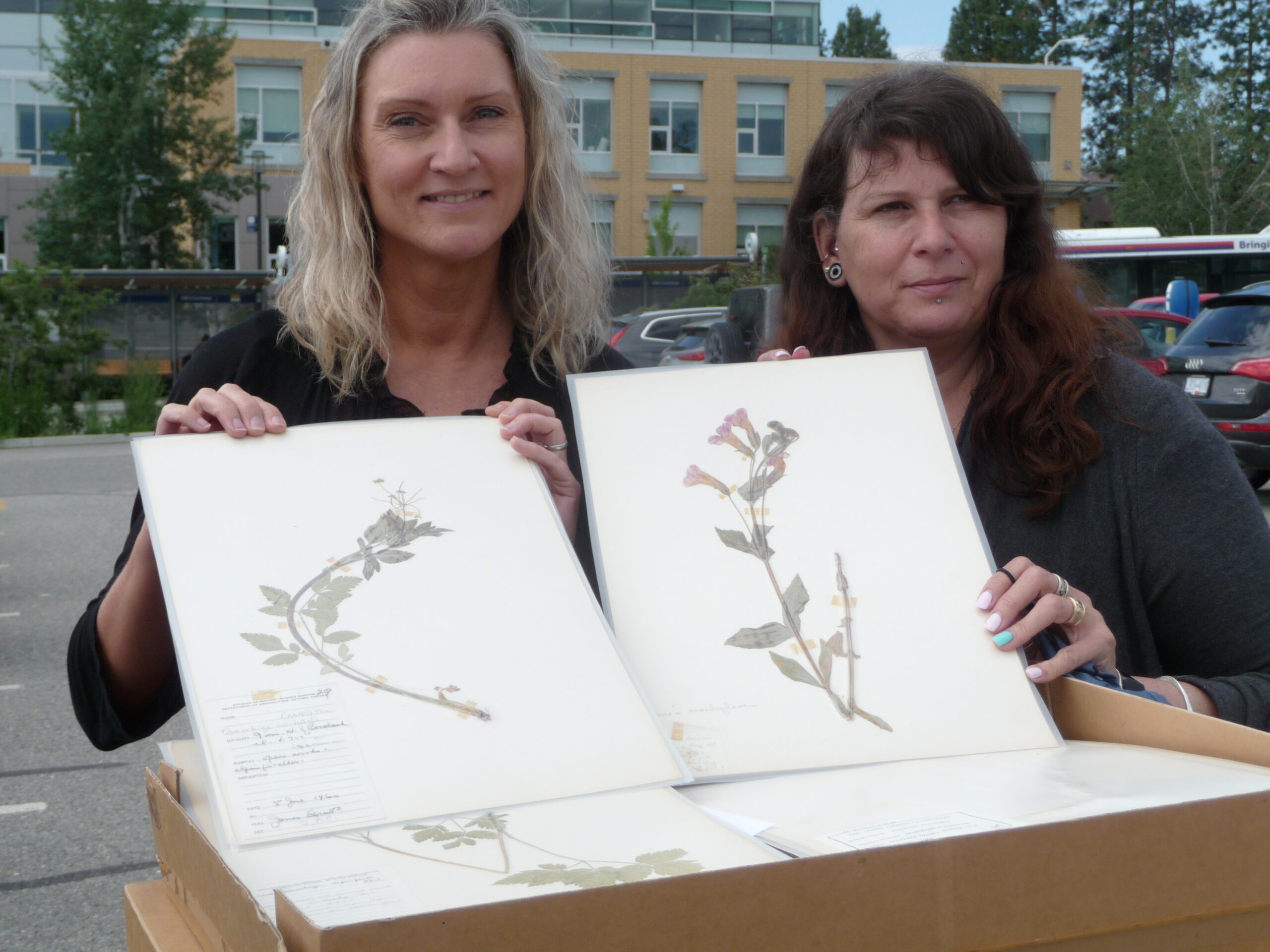 Two women in front of a beige, multi-storied building, holding botanical samples. The woman on the left is blond, and smiling at the camera. The woman on the right has dark red hair with bangs, with piercings on her hair and one below her mouth, and smiling slightly and looking off to her left. The left sample is a green plant with a white flower pressed flat on a sheet of paper between sheets of thin plastic. The right sample has pink, bell-shaped flowers. 