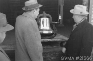 A black and white image of a large metallic bell on a table with three men looking at it. All three are wearing bowler hats, and have their backs to the camera. The bell is hanging in a metal sling contraption.