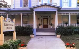 a paved pathway leads to an entry way of a blue house with white trim under an arched verandah. To the left, a large, free-standing sign, painted yellow, reads "Sultenfuss Parlor."