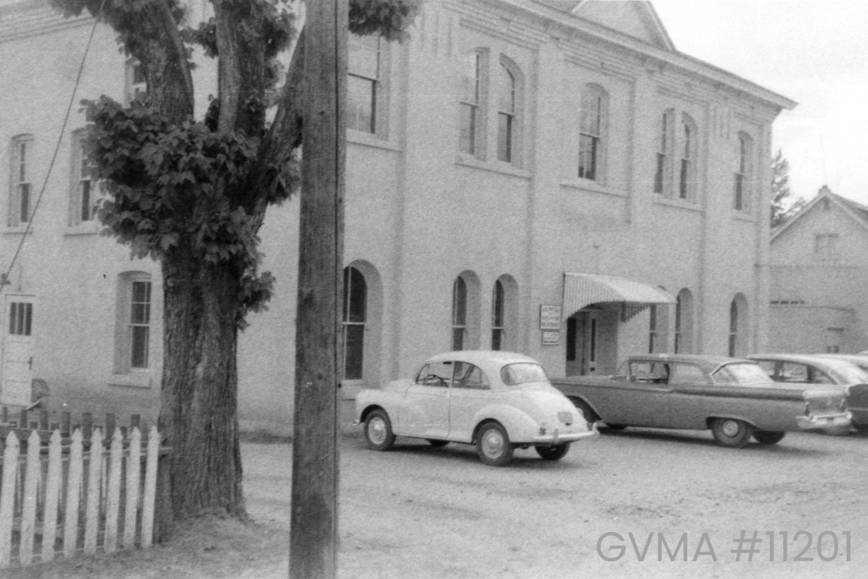 A slightly-angled view of a black and white school building with arched windows and a curtain verandah. It has two stories. Several old-fashioned cars are parked in the front.