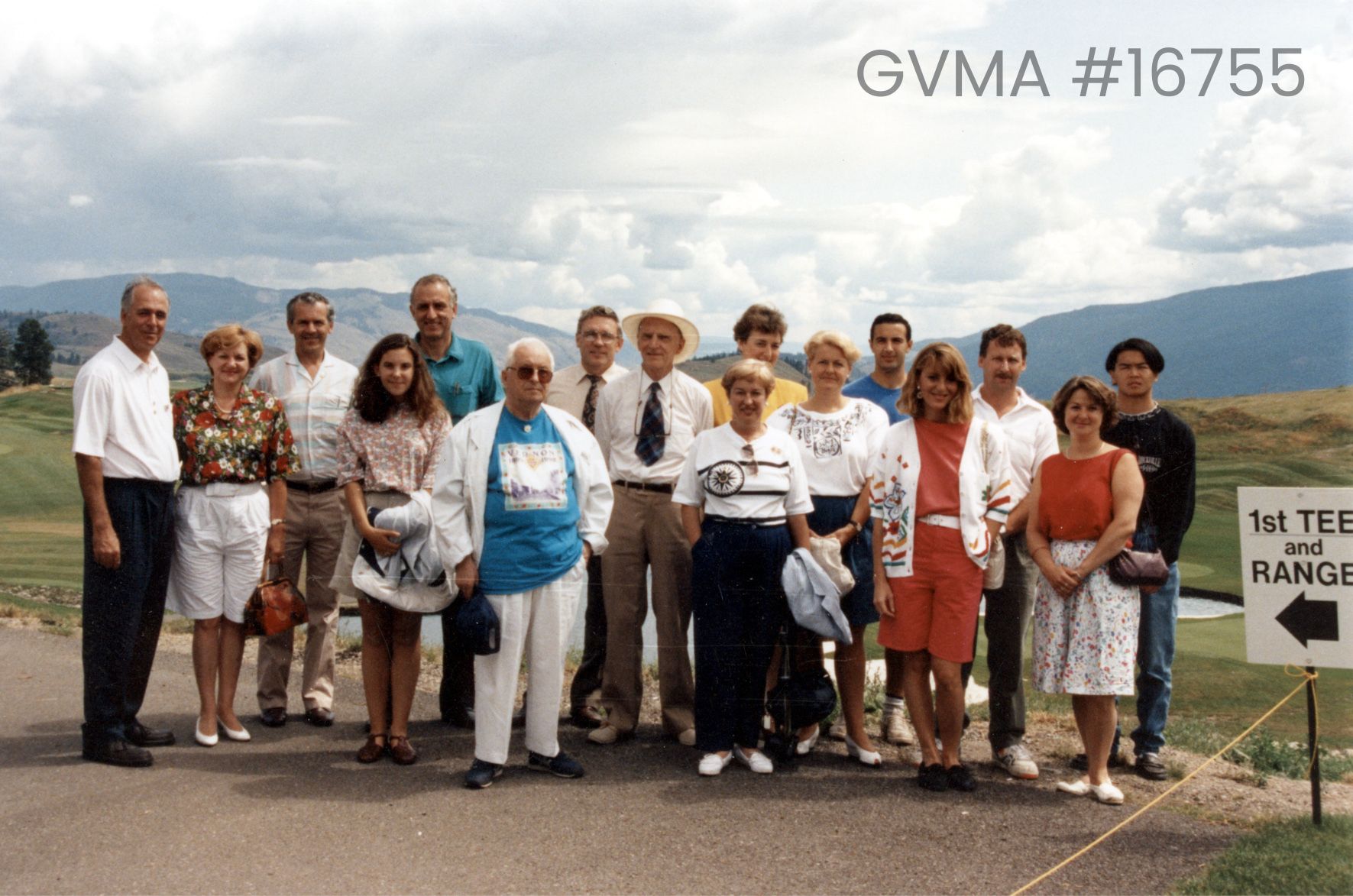 a group of men and women standing facing the camera. Behind them is a golf course, and on the right is part of a sign that reads "1st tee and range" with an arrow.