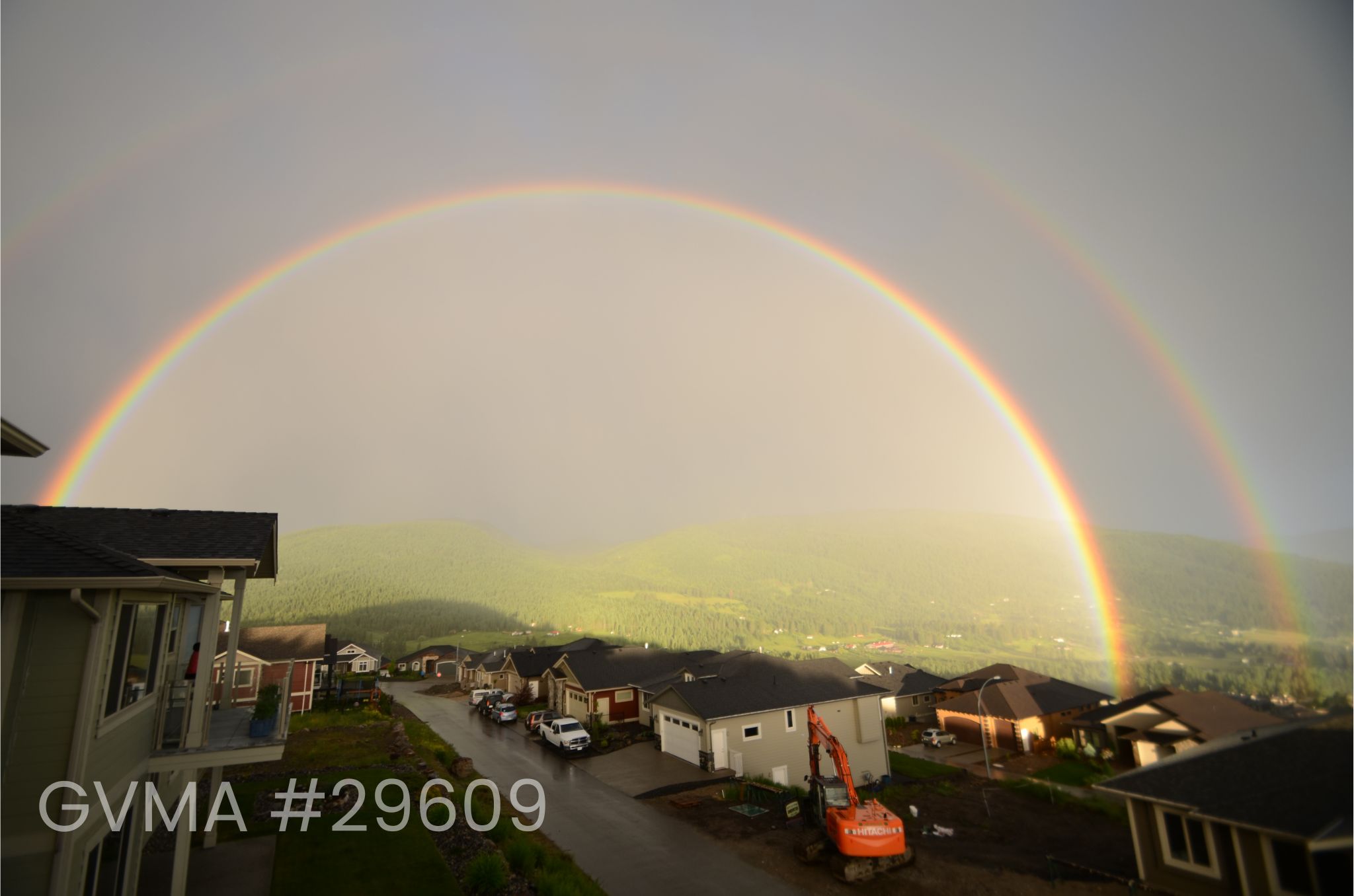 A view down a street over the rooftops of houses. A green hillside is visible in the background. A double rainbow is also visible, with the bottom one being much stronger and the top one quite faint.