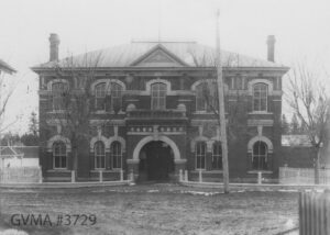 A front view of a black and white building white a dark brown brick exterior and white trim. The doorways and windows are arched at the top. Several spindly tress without leaves are standing in front of the two-story building and an unpaved road is in front of it.