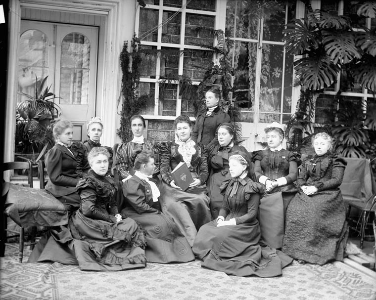 A black and white photo of 11 women, some seated on chairs, some in front on the floor, with one standing in the back. They are in a room lined with bookshelves that has brick walls and several large houseplants/.