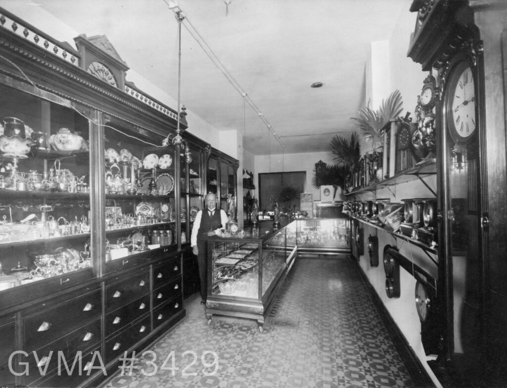 A man is standing in a long room and leaning on a glass display case. All along the walls are tall wooden cabinets filled with silverware and porcelain.