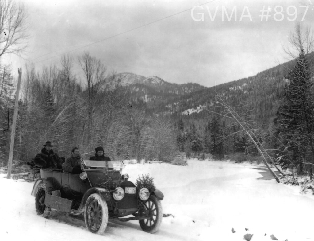 A black and white image of an old car with three occupants and some greenery attached to it on a snowy backroad. In the background is a mountain and trees. 