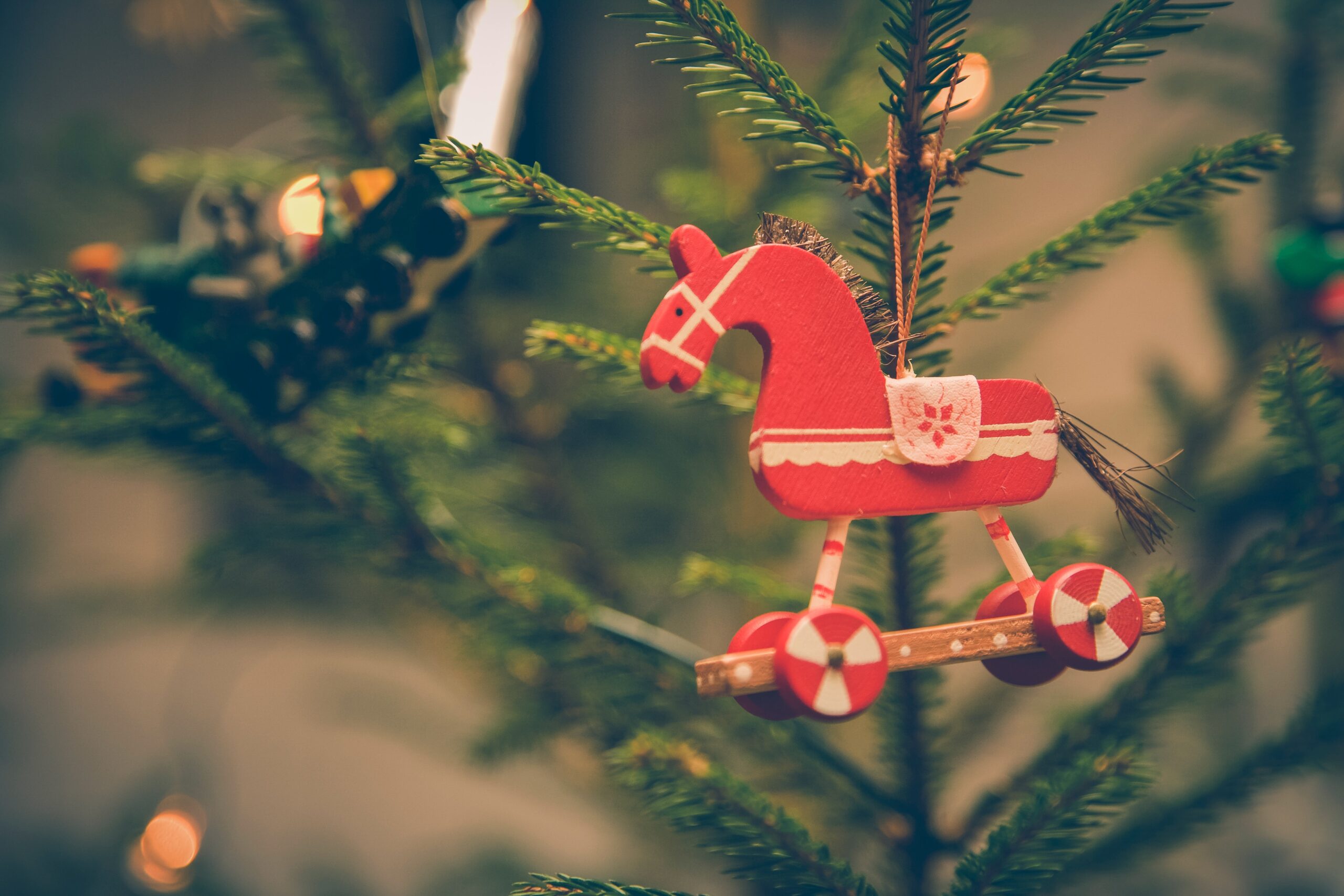 A close-up of a green Christmas Tree with lights. A small red wooden rocking horse decoration in hanging in the foreground.