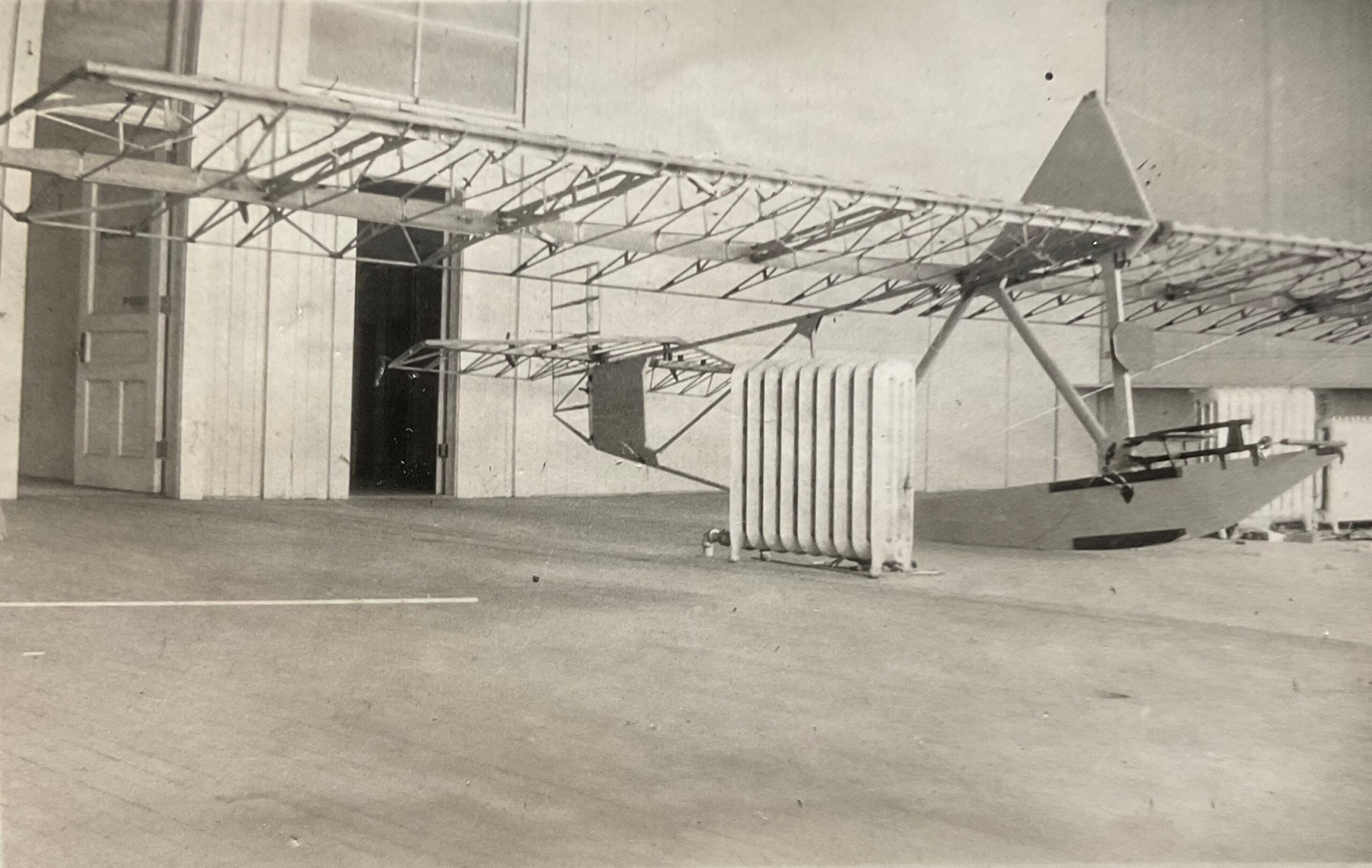 A large frame of a glider under construction in an empty room. A space heater is nearby.