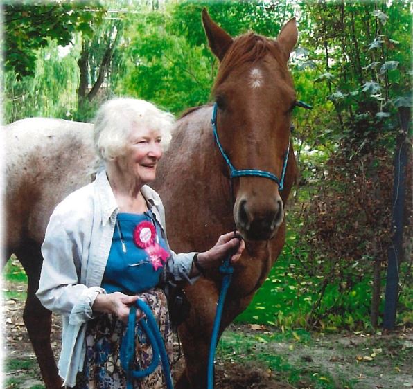 A woman with grey hair wearing a jean shirt over a blue patterned dress has a pink birthday badge pinned to her dress. She is outdoors, and leading a horse by a rope.