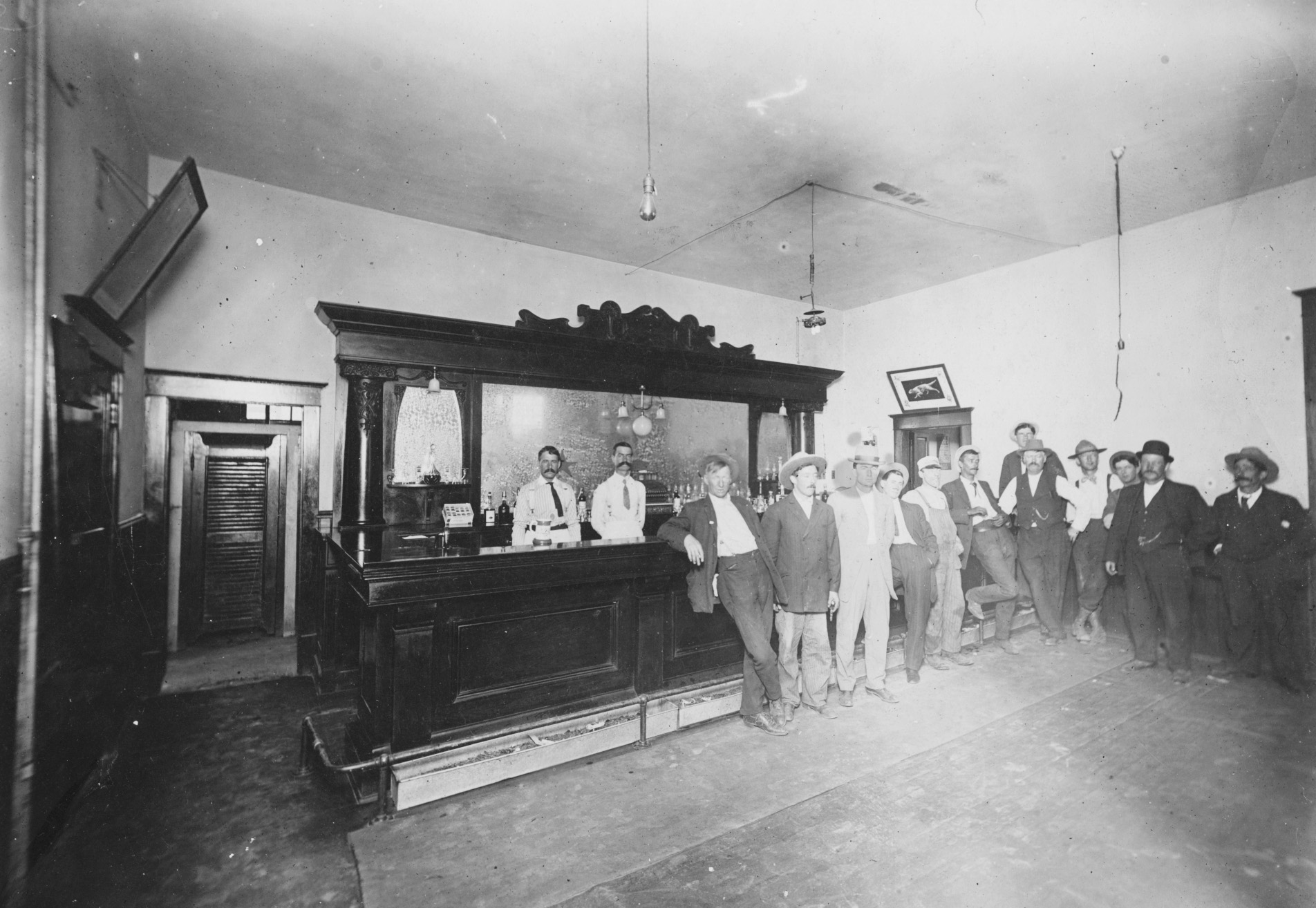 A black and white image of a large bar room. A dark bar is set against the far wall, and three men in white uniforms are standing behind it. A number of men are standing in front of the bar.