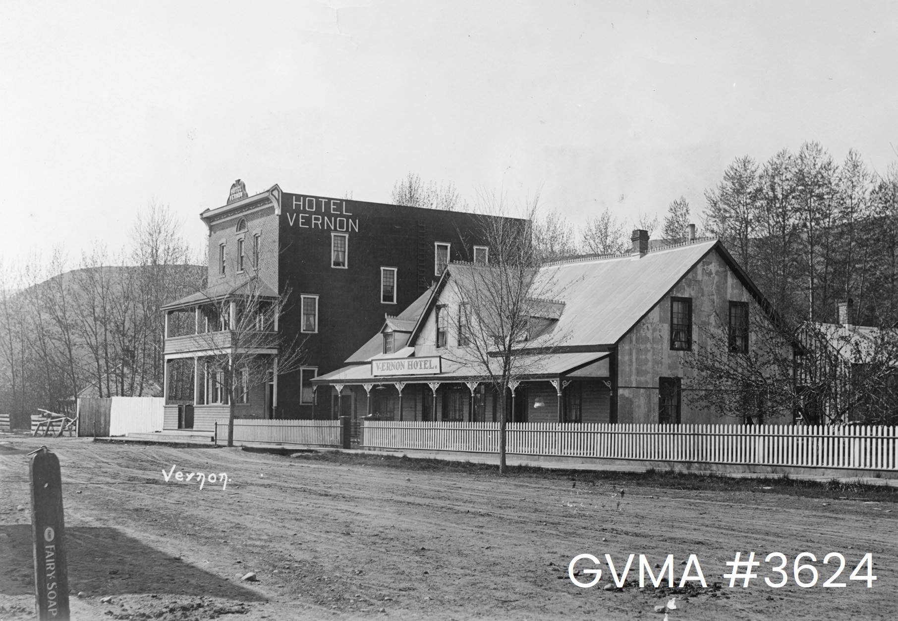 A black and white image of two buildings on the side a dirt road. The structure in the foreground, the Vernon Hotel, is shorter while the Hotel Vernon next to it is three-stories tall.
