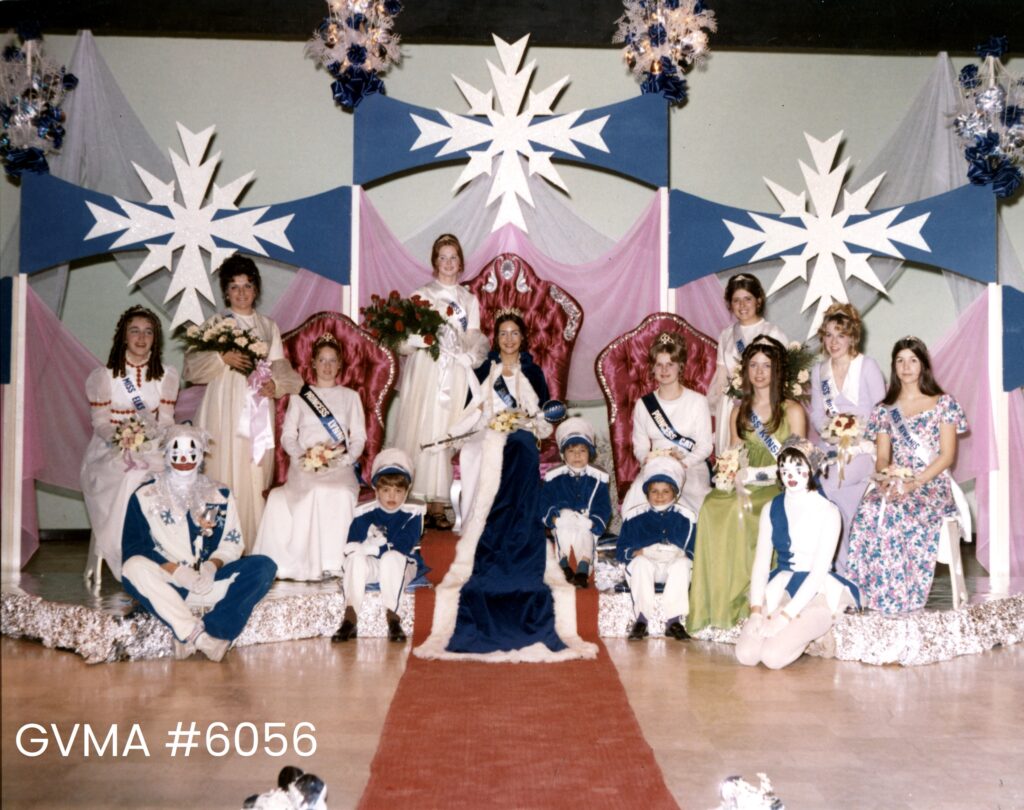 A group of young women in dresses, a blue-and-white jester, and three young pages seated on chairs beneath blue banners with large white snowflakes.