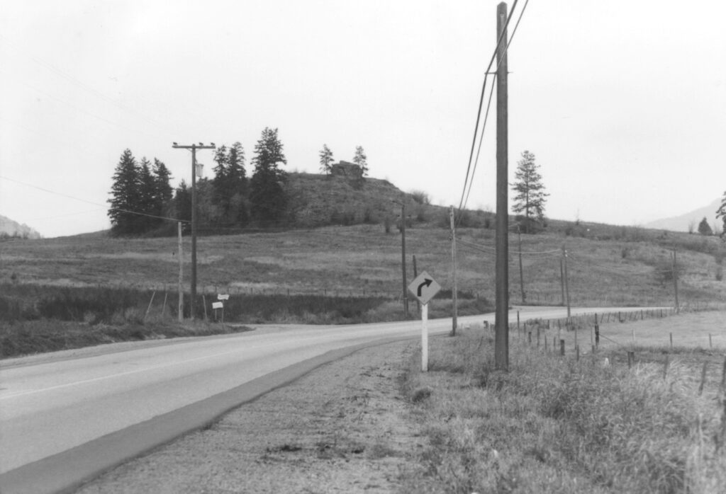 A black and white image of a large boulder sitting on the top of a distant hill. In the foreground is a highway,