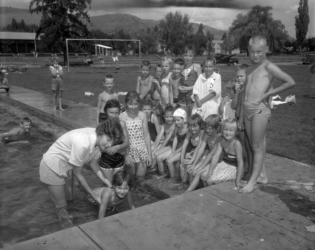 A black and white image of children standing around a pool, with some dangling their feet in the water, and one child swimming while being held on to by an instructor.