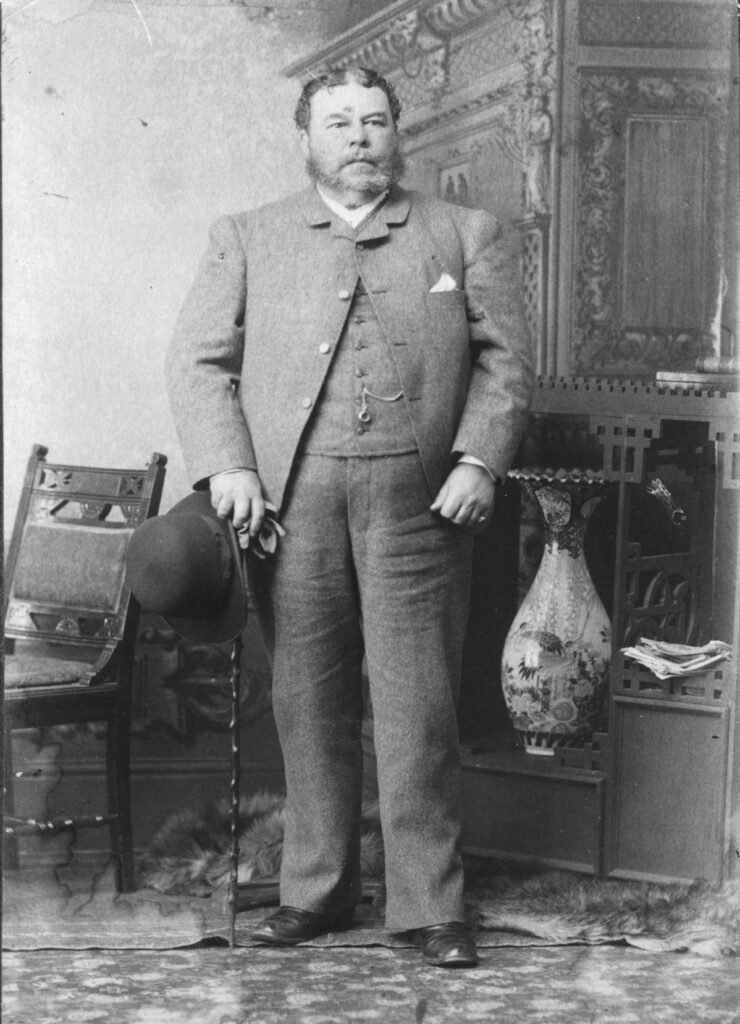 A black-and-white photo of a man standing  in a room. He has an ornate cane and a hand in one hand. He is wearing a suit and pocket watch.