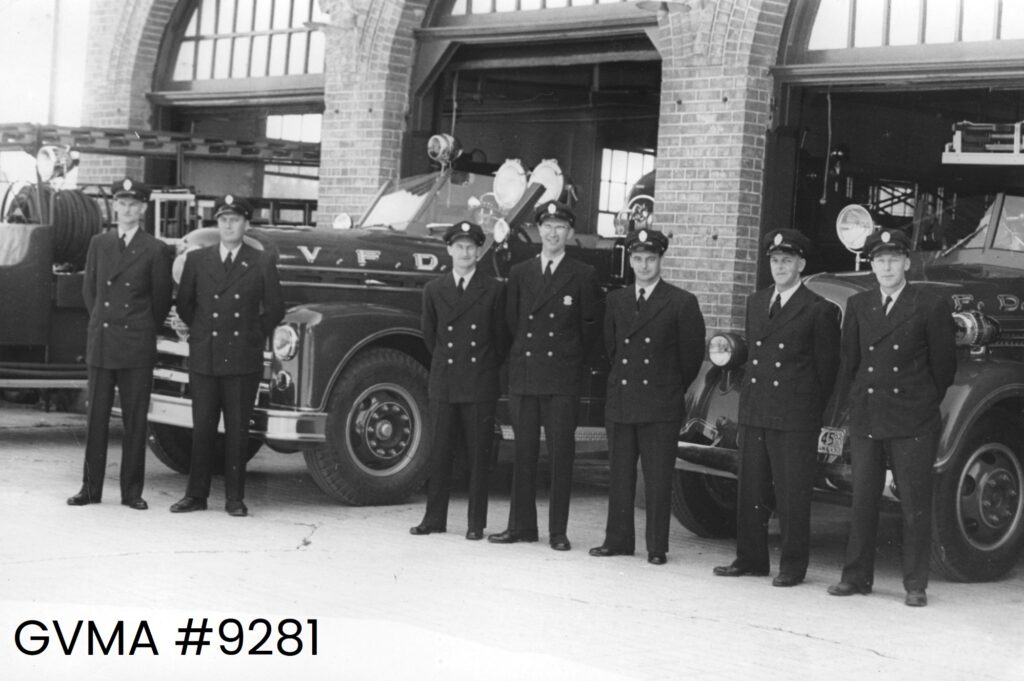 A black-and-white image of a group of men standing outside a fire hall. There are 7 men pictured and three trunks behind them.