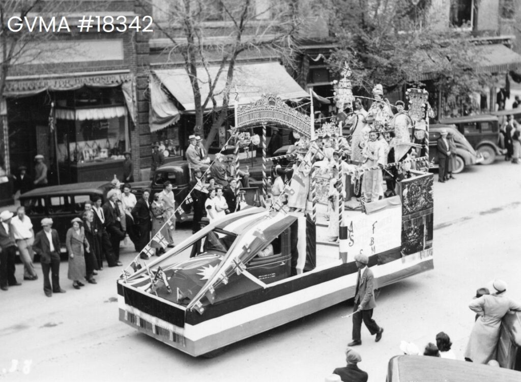 A black and white image of a float moving through Vernon's main street. The float is decorated with union jacks and chinese regalia and several women and girls in traditional dress are seated on the back.