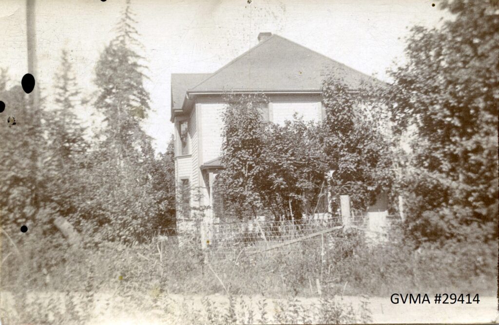 A sepia image of a multi-story house, taken from the side, and mostly concealed by trees and shrubs.