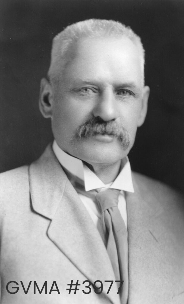 A black and white image of a man in a white shirt with a pale suit jacket and tie. He has a handle bar mustache and short, white hair.