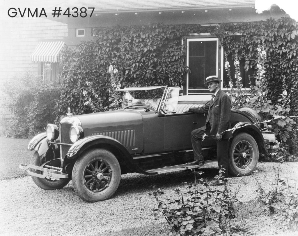 Black and white image of a roofless car in front of a building covered in foliage. A man is standing next to it, with one leg resting on the bumber.