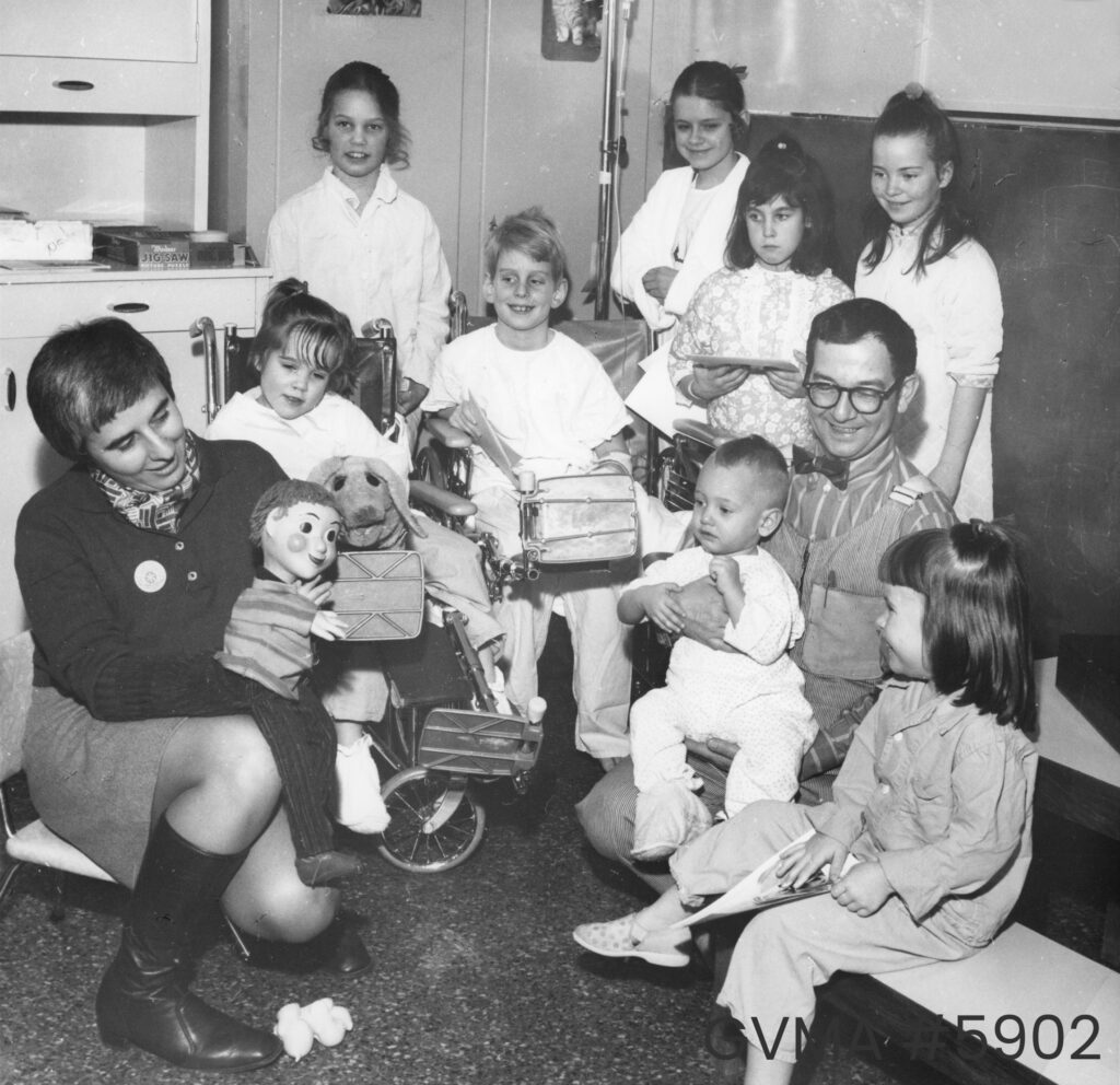 Black and white image of a man and woman with eight children of various agees around them, with a few of them in wheelchairs. Ernie Coombs is sitting with an infant on his lap, and Judith Lawrence is kneeling and holding two puppets.