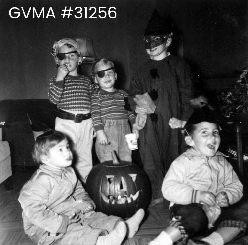 a black-and-white image of five children. Three are standing and two are sitting around a carved pumpkin. Two are wearing eyepatches and one is dressed as a clown. The two seated are wearing part hats.