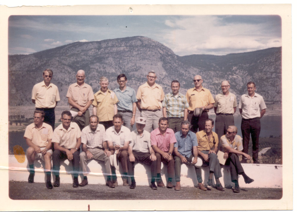 Colour photo of a group of horticulturalists posing at the side of a road in front of a lack. 18 men are pictured. Peter Humphry-Baker is pictured wearing a blue shirt.