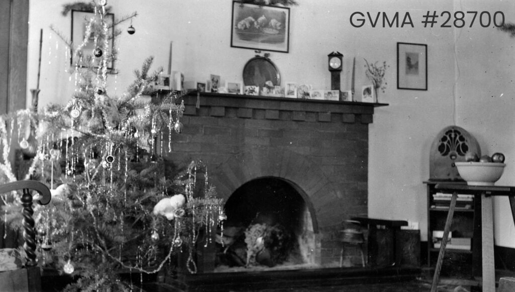 A black-and-white image of a fireplace next to a medium-sized tree decorated with tinsel. A few pieces of pottery line the top of the brick fireplace.