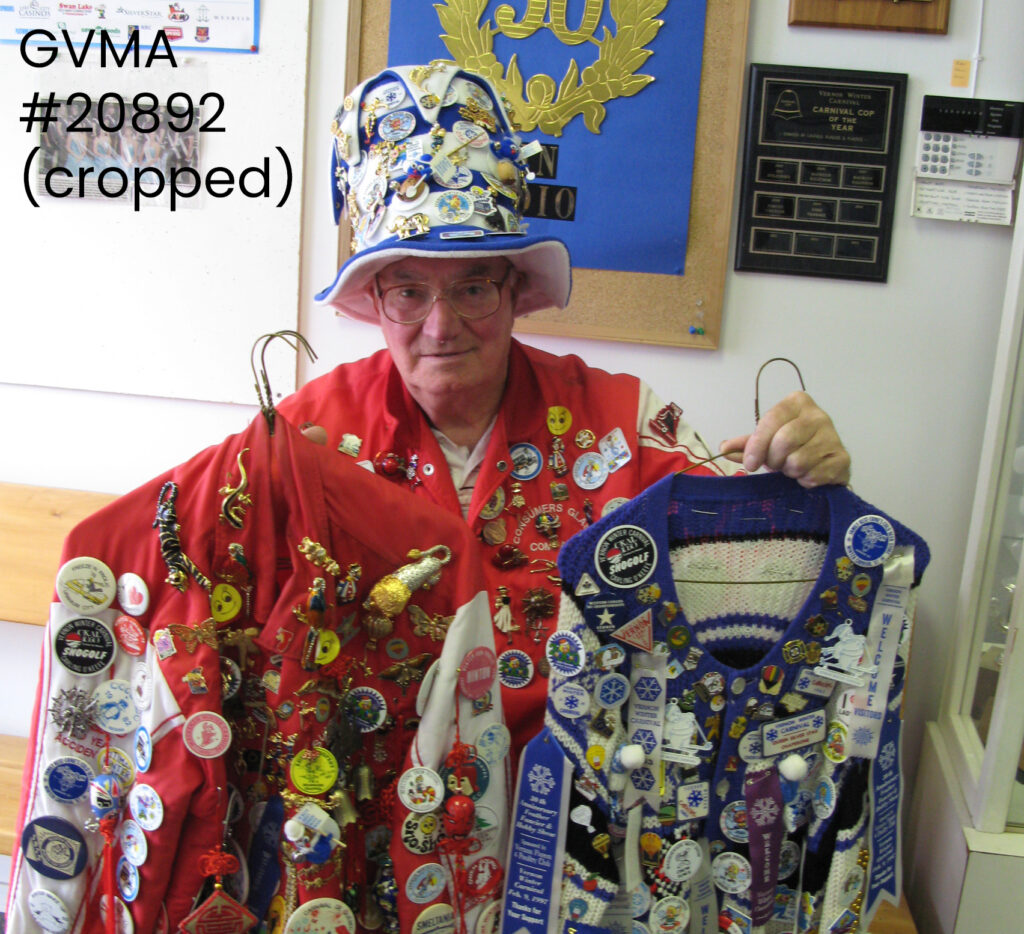 A man wearing a red bomber jacket and white and blue top hat holding another red jacket and a blue and white jacket. All of the jackets and the hat are covered in pins and buttons.