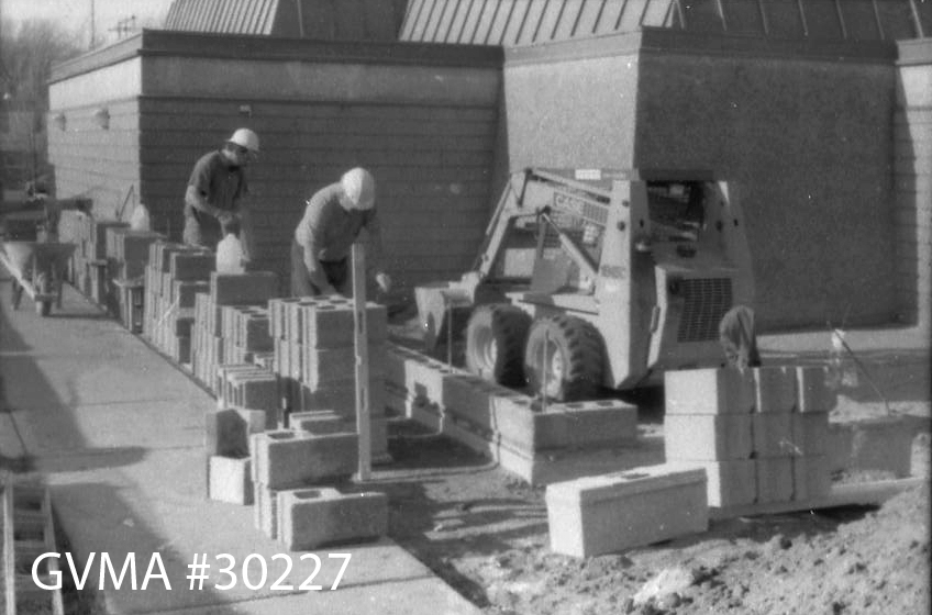 A black and white image showing two men in hard hats next to a pile of bricks outside a concrete building. Beside them is a piece of heavy machinery.